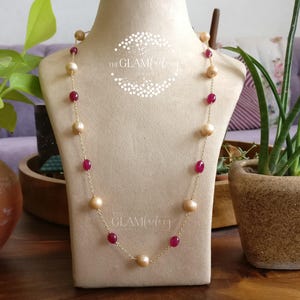 Op de afbeelding: Een gouden ketting met afwisselend grote, lichtgekleurde parels en kleine, ovale, rode edelstenen. De ketting wordt getoond op een beige mannequin. Het logo van The Glam Factory is zichtbaar.