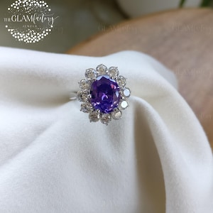 May include: A silver ring featuring a large, oval-cut purple gemstone surrounded by a halo of smaller, clear stones. The ring is set on a white fabric background, showcasing the jewelry's sparkle and design. The Glam Factory logo is in the upper left corner.