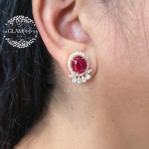 May include: Gold-toned stud earrings featuring a large, oval-shaped, deep red gemstone surrounded by a halo of small, clear stones. Below the main stone are three smaller, clear, marquise-cut stones. The earrings are set against a skin tone background.