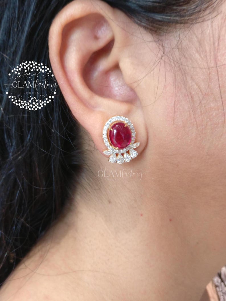 May include: Gold-toned stud earrings featuring a large, oval-shaped, deep red gemstone surrounded by a halo of small, clear stones. Below the main stone are three smaller, clear, marquise-cut stones. The earrings are set against a skin tone background.