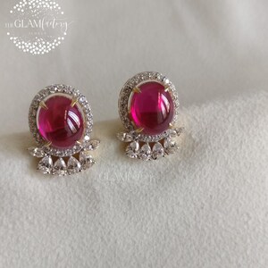 May include: A pair of stud earrings featuring large, round, magenta-colored gemstones surrounded by a halo of small, clear crystals. The earrings have a gold-colored setting and a cluster of teardrop-shaped crystals below the main stone.