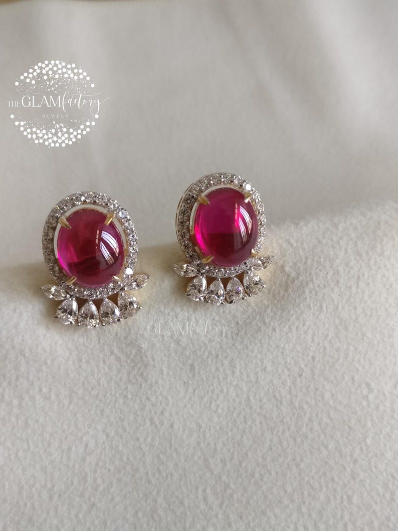 May include: A pair of stud earrings featuring large, round, magenta-colored gemstones surrounded by a halo of small, clear crystals. The earrings have a gold-colored setting and a cluster of teardrop-shaped crystals below the main stone.