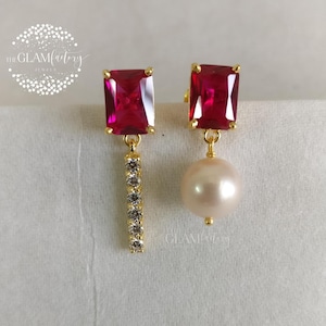Peut inclure: Paire de boucles d'oreilles asymétriques. L'une présente une pierre carrée rouge rubis avec une barre de pierres claires. L'autre a une pierre carrée rouge rubis et une perle. Le logo The Glamfactory est visible.