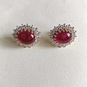 May include: A pair of stud earrings featuring large, oval, deep red gemstones surrounded by a halo of small, clear, sparkling stones. The earrings have a gold-colored setting, creating a luxurious and elegant appearance.