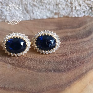 Puede incluir: Un par de pendientes de oro con una gran piedra preciosa ovalada azul rodeada por un halo de piedras preciosas m&aacute;s peque&ntilde;as y transparentes.