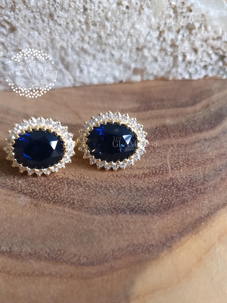 Puede incluir: Un par de pendientes de oro con una gran piedra preciosa ovalada azul rodeada por un halo de piedras preciosas m&aacute;s peque&ntilde;as y transparentes.