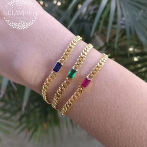 May include: Gold-coloured chain bracelets, each with a rectangular gemstone in blue, green, and pink. The bracelets are shown on a wrist, highlighting the jewellery's design and colour variations. The Glam Factory logo is visible in the background.