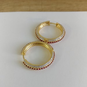 May include: A pair of gold hoop earrings with a double row of clear cubic zirconia stones and a single row of red cubic zirconia stones.