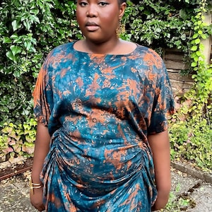 May include: A midi-length dress featuring a teal and copper-brown abstract marbled pattern. The garment has a round neckline, short sleeves, and gathered side detailing that creates a draped effect along the waist and hips.
