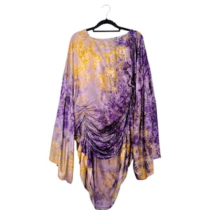 May include: A long-sleeved tunic top featuring a marbled tie-dye pattern in shades of purple and gold. The garment has a round neckline and a gathered, ruched detail along the left side that creates an asymmetrical hemline and draped effect.