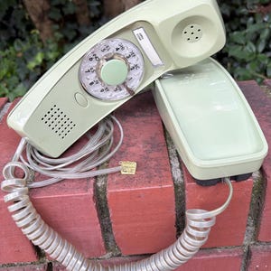 May include: A light green wall-mounted telephone with a rotary dial featuring letters and numbers. The handset rests on the base unit, connected by a clear coiled cord with a modular plug. A small rectangular label area is visible above the dial.