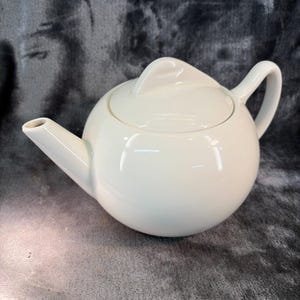 May include: A glossy off-white ceramic teapot with a spherical body, a curved handle, a tapered spout, and a matching lid featuring a triangular handle. The teapot has a smooth, reflective surface and a clean, rounded silhouette throughout.