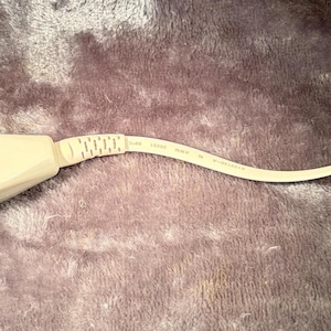 May include: A beige EWIRE branded adapter featuring a rectangular housing with a textured grip section, connected to a short, flat cable that terminates in a clear plastic RJ11 telephone connector. The device is designed for modular connectivity.