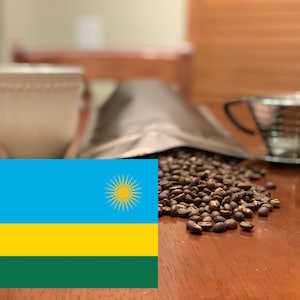 May include: A still-life image featuring coffee-related items. A white mug, a silver coffee dripper, and a bag of coffee beans are arranged on a wooden surface. The image also includes a pile of roasted coffee beans and the flag of Rwanda.