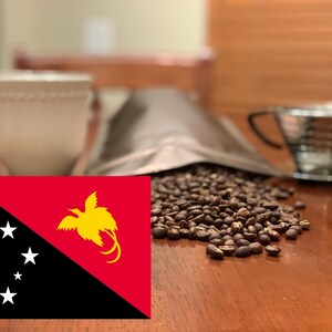 May include: A still life featuring coffee beans, a white mug, a metal pour-over coffee maker, and a bag of coffee beans. The flag of Papua New Guinea is in the foreground. The coffee beans are scattered on a wooden surface.