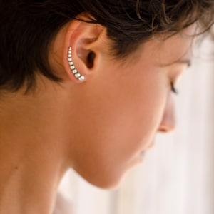 Puede incluir: Un pendiente de aro de plata con una fila de pequeñas cuentas redondas. El aro es curvo y se coloca en la parte superior de la oreja.