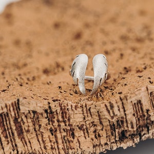May include: A silver-colored ring with an open design, featuring two stylized wing shapes. The ring is set against a textured, brown surface, creating a contrast in color and texture. The wings have a detailed, feathered appearance.