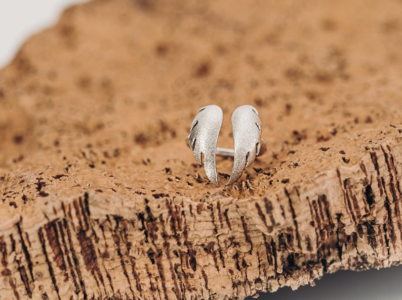 May include: A silver-colored ring with an open design, featuring two stylized wing shapes. The ring is set against a textured, brown surface, creating a contrast in color and texture. The wings have a detailed, feathered appearance.