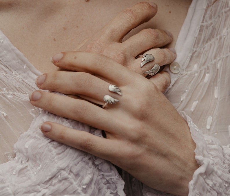 May include: Silver rings with wing designs. One ring features two wings, while another has a single wing design. The rings are worn on fingers, with the hands resting together. The rings are a unique jewelry item.