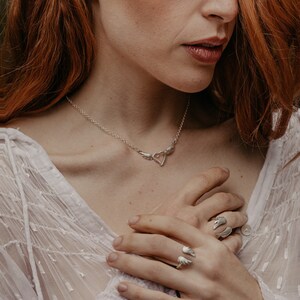 May include: A silver jewelry set featuring a necklace with a heart pendant and wing accents, matching wing earrings, and two wing-shaped rings. The jewelry is worn on a person with red hair and a white top.