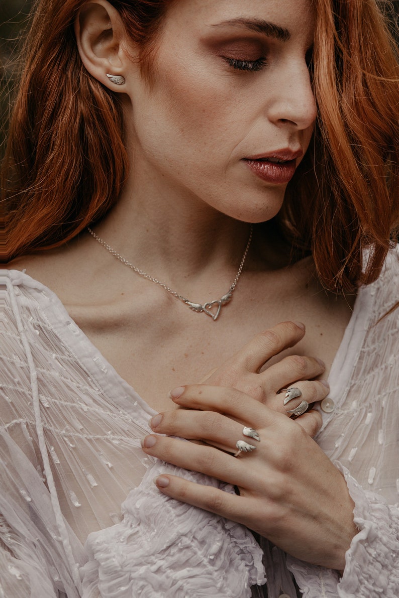 May include: A silver jewelry set featuring a necklace with a heart pendant and wing accents, matching wing earrings, and two wing-shaped rings. The jewelry is worn on a person with red hair and a white top.