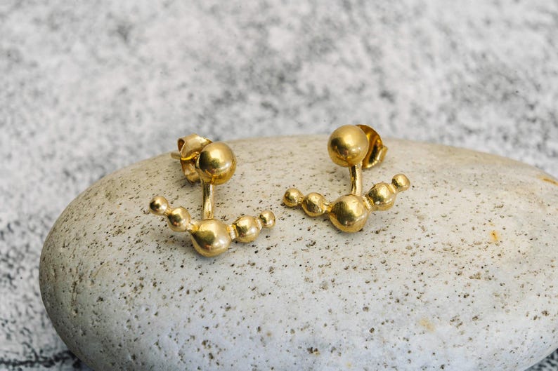 May include: A pair of gold-toned earrings with a curved design featuring multiple small spheres. The earrings are designed to be worn on the upper earlobe.