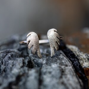 May include: A silver-colored ring featuring an open design with two textured wing shapes. The ring is set against a blurred background of a gray, textured stone. The wings are detailed with feather-like patterns.