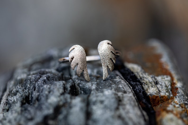 May include: A silver-colored ring featuring an open design with two textured wing shapes. The ring is set against a blurred background of a gray, textured stone. The wings are detailed with feather-like patterns.