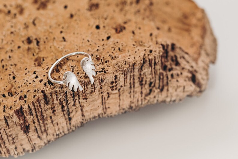 May include: A silver-colored ring with a leaf design. The ring features two open-ended leaf shapes. The ring is displayed on a textured, brown surface.