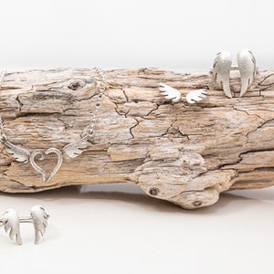 May include: A collection of silver-toned jewelry featuring a heart and wing design. The set includes a necklace, a ring, a pair of earrings, and a ring with wing accents. The jewelry is displayed on a piece of driftwood against a white background.