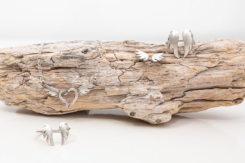 May include: A collection of silver-toned jewelry featuring a heart and wing design. The set includes a necklace, a ring, a pair of earrings, and a ring with wing accents. The jewelry is displayed on a piece of driftwood against a white background.