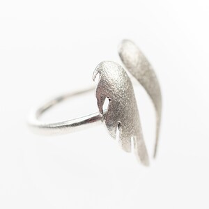 May include: A silver-colored ring with a unique wing design. The ring features two textured, feather-like elements that curve upwards. The ring is set against a white background, highlighting its metallic sheen. A stylish piece of jewelry.