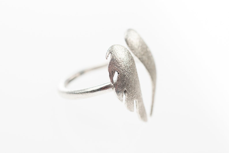 May include: A silver-colored ring with a unique wing design. The ring features two textured, feather-like elements that curve upwards. The ring is set against a white background, highlighting its metallic sheen. A stylish piece of jewelry.