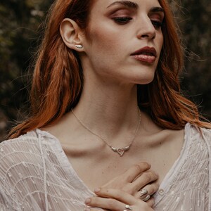 May include: A silver necklace, ring, and earrings set. The necklace features a heart pendant with wing accents. The ring and earrings also have wing designs. The jewelry is worn with a white, sheer top.