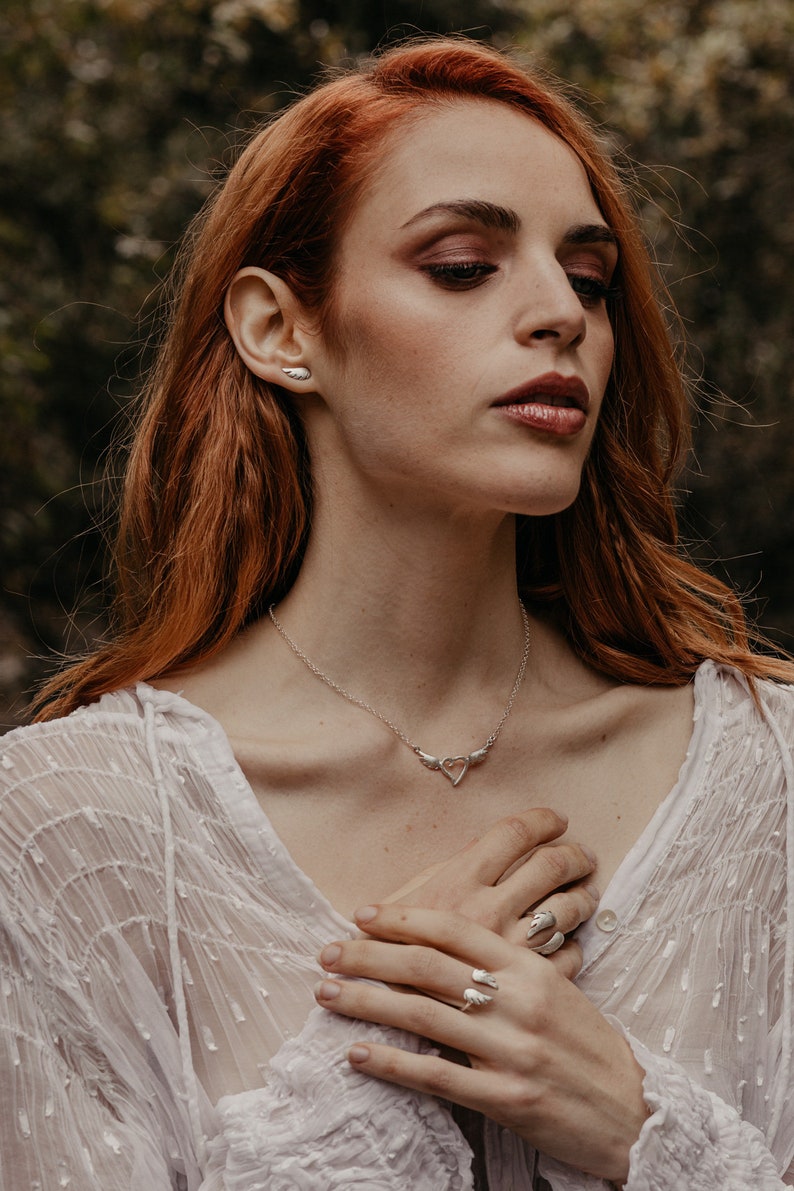 May include: A silver necklace, ring, and earrings set. The necklace features a heart pendant with wing accents. The ring and earrings also have wing designs. The jewelry is worn with a white, sheer top.
