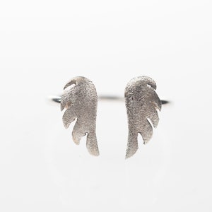 May include: A silver ring featuring a pair of textured angel wings. The wings are detailed with feather-like patterns and are positioned to face outward. The ring is set against a white background.