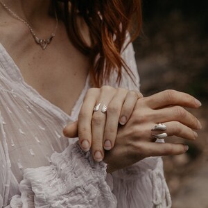 May include: A close-up of two silver rings and a necklace. One ring features an open wing design, while the other has a curved, layered shape. The necklace has a delicate, symmetrical pendant. The jewelry is worn on a person's hand.