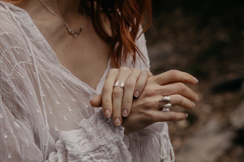 May include: A close-up of two silver rings and a necklace. One ring features an open wing design, while the other has a curved, layered shape. The necklace has a delicate, symmetrical pendant. The jewelry is worn on a person's hand.
