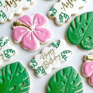 May include: A set of nine decorated sugar cookies with a tropical theme. The cookies are decorated with pink hibiscus flowers, green monstera leaves, and gold glitter. One cookie has the words "Happy Birthday" written on it. Another cookie has the name "Anya" written on it.
