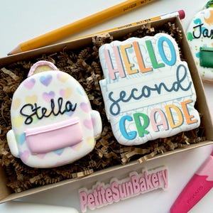 May include: A box of decorated sugar cookies shaped like backpacks and school-themed designs. One cookie says "Hello Second Grade", another says "Stella", and a third says "Jaxson". A pink pair of scissors and a pencil are also in the image.