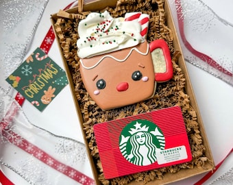 Könnte beinhalten: Ein Lebkuchen-Tassenkeks mit weißem Zuckerguss, Zuckerstange und Streuseln, in einer Pappschachtel mit einer Starbucks-Geschenkkarte. Ein kleines Weihnachtsetikett mit "Merry Christmas" ist ebenfalls enthalten.