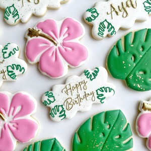 May include: A set of decorated sugar cookies with a tropical theme. The cookies feature pink hibiscus flowers, green monstera leaves, and white cookies with the words "Happy Birthday" and "Aya" in gold lettering.