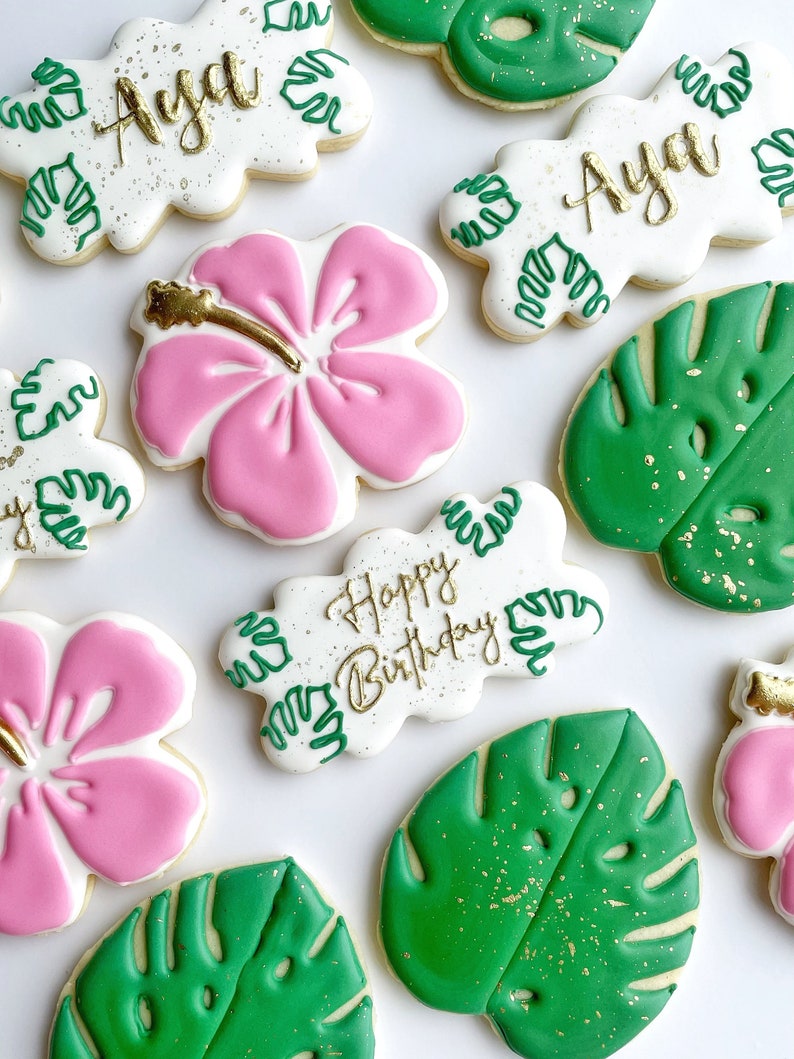 May include: A set of decorated sugar cookies with a tropical theme. The cookies feature pink hibiscus flowers, green monstera leaves, and white cookies with the words "Happy Birthday" and "Aya" in gold lettering.