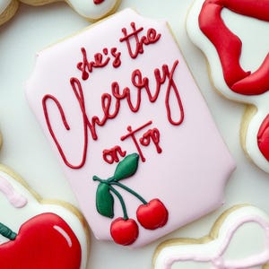 Pu&ograve; includere: Un biscotto rettangolare con angoli arrotondati, ricoperto da una glassa rosa chiaro. Il testo 'she's the Cherry on top' &egrave; scritto in glassa rossa, accompagnato da due ciliegie rosse con foglie verdi nella parte inferiore.