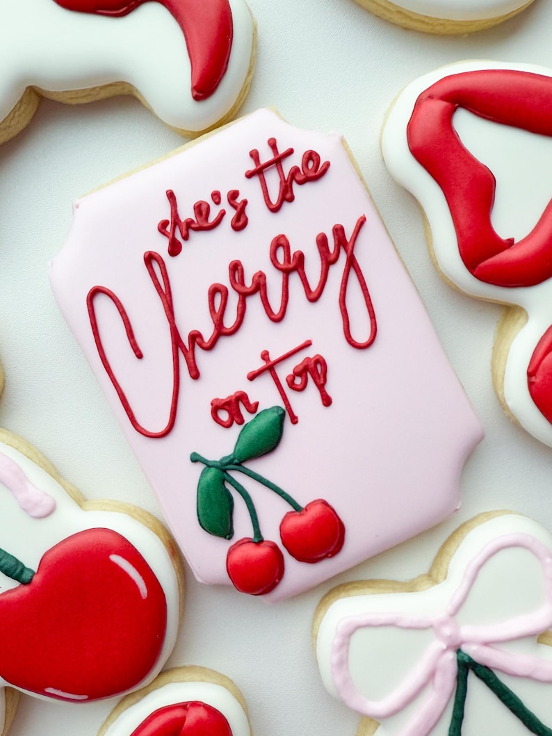 Pu&ograve; includere: Un biscotto rettangolare con angoli arrotondati, ricoperto da una glassa rosa chiaro. Il testo 'she's the Cherry on top' &egrave; scritto in glassa rossa, accompagnato da due ciliegie rosse con foglie verdi nella parte inferiore.