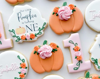 Puede incluir: Una variedad de galletas de azúcar decoradas. Las galletas incluyen formas de calabaza naranja cubiertas con glaseado rosa, formas del número uno y galletas redondas con el nombre "Isabella". Algunas galletas tienen detalles florales y la frase "Our Little Pumpkin is Turning ONE".