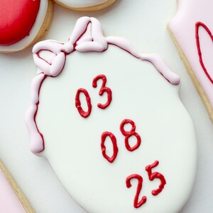 Pu&ograve; includere: Un biscotto bianco di forma ovale decorato con i numeri 03, 08 e 25 in glassa rossa impilati verticalmente. Il biscotto presenta un fiocco in glassa rosa chiaro sulla parte superiore e un bordo rosa chiaro lungo i lati della superficie.