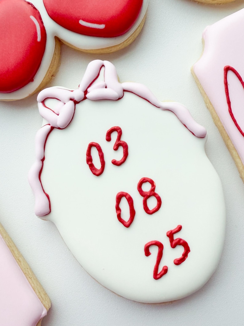 Pu&ograve; includere: Un biscotto bianco di forma ovale decorato con i numeri 03, 08 e 25 in glassa rossa impilati verticalmente. Il biscotto presenta un fiocco in glassa rosa chiaro sulla parte superiore e un bordo rosa chiaro lungo i lati della superficie.