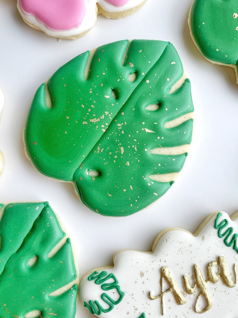 May include: Green sugar cookies decorated with green icing and gold sprinkles, shaped like monstera leaves.
