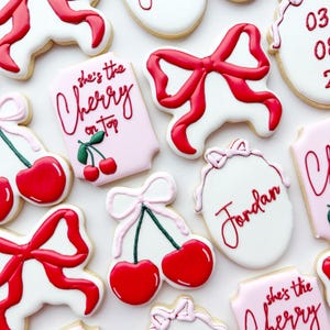 Pu&ograve; includere: Una collezione di biscotti decorati con fiocchi di glassa rossa, targhette rettangolari rosa e bianche con il testo 'she's the Cherry on top', biscotti ovali con il nome 'Jordan' o le date '03 08' in rosso e motivi a ciliegia con fiocchi rosa.